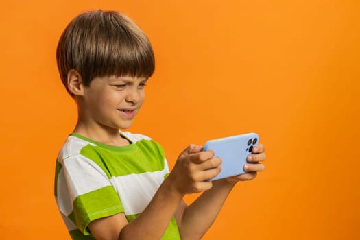 Happy excited young preteen child boy kid player celebrate victory enthusiastically playing racing drive simulator, shooter video games on smartphone. Little children isolated on orange background