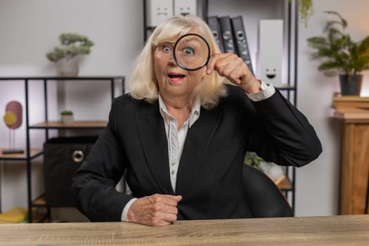 Investigator researcher businesswoman looking through magnifying glass with wow expression with big zoomed funny eye, searching, analyzing. Professional Caucasian freelancer grandma looking at camera.