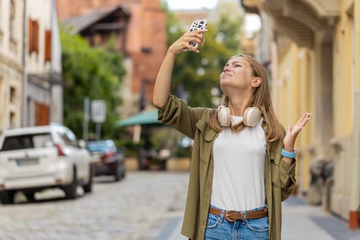Young female tourist woman trying to catch a communication signal and Internet on mobile phone, raised the smartphone up for a better connection in city street. Frustrated Caucasian girl in town.