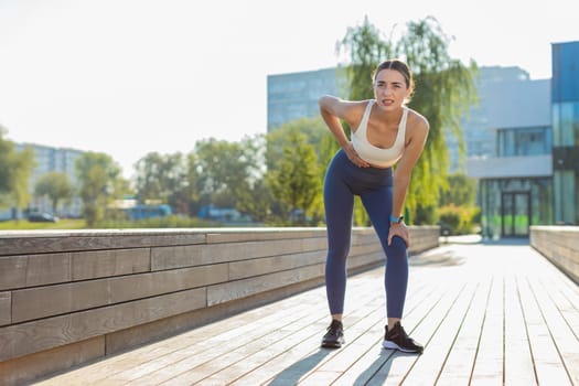 Young Caucasian runner suffering from abdominal pain during a run in urban city park on sunny day. Sad woman keeps her hand on her stomach. Girl has poisoning, constipation, spasms or menstruation.