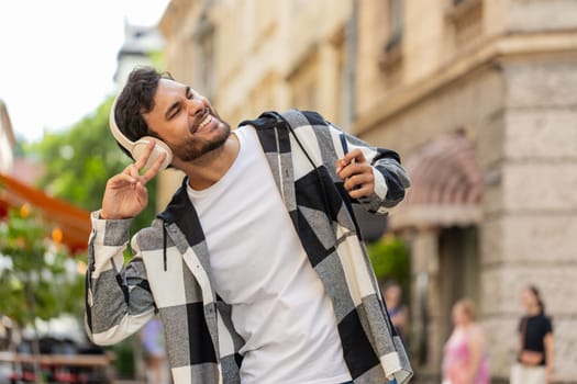 Happy overjoyed Arabian young man tourist in wireless headphones choosing, listening favorite energetic disco music on smartphone outdoors. Indian tourist guy walking passes by urban city street.