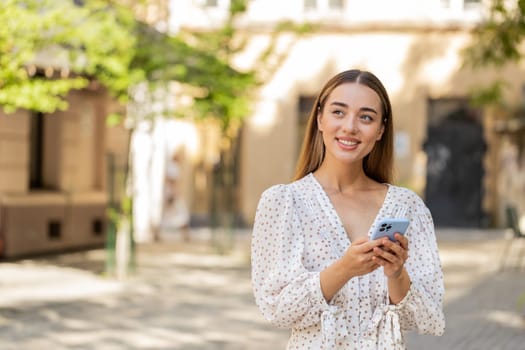 Caucasian young woman in long dress using smartphone typing texting social media messages e-mail looking for a way on map in mobile navigator app outdoors. Girl standing in city street. Town lifestyle