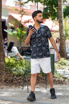 Tourist traveler young bearded man using smartphone typing text messages looking for search a way on map in mobile navigator app outdoors. Indian guy male walking passes by urban city street park.