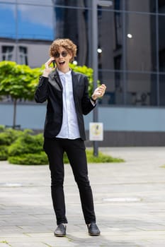 Caucasian businessman making phone call online conversation, mobile loudspeaker talking, waving hello outside office workplace. Young man in formal suit with curly hair on video call via smartphone.