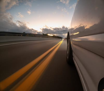 A car is driving down a road with a sunset in the background. The car is white and has its headlights on