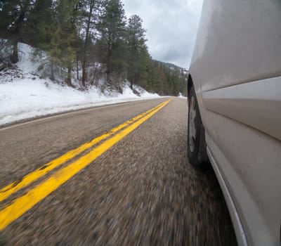 A car is driving down a snowy road with yellow lines. The car is white and has a tire that is visible. The scene is blurry, giving the impression of motion