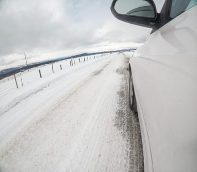 A car is driving down a snowy road with a fence in the background. The car is in the driver's mirror and the road is covered in snow