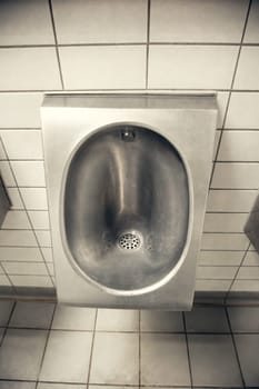 A stainless steel urinal installed in a public restroom with white tiled walls. Minimalist and industrial style, emphasizing hygiene, cleanliness, and functional design