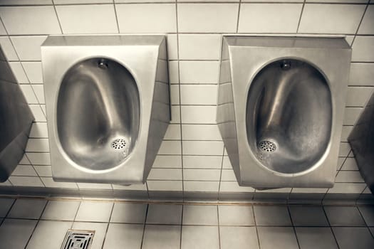 Stainless steel urinals in a public restroom with white tiled walls. The industrial and functional design is perfect for hygiene-related themes, public facilities, and restroom interiors