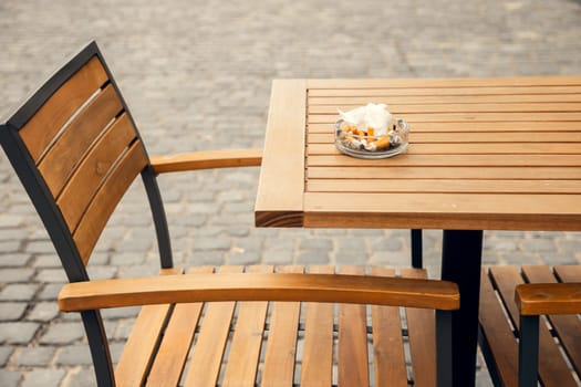 A wooden outdoor cafe table with a full ashtray and a matching chair on a cobblestone pavement. A realistic depiction of outdoor dining culture, smoking habits, and urban life