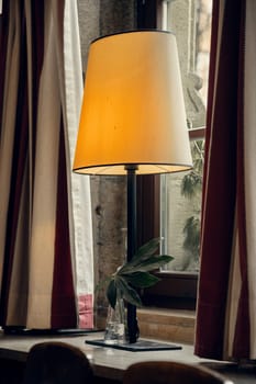 A warmly lit table lamp standing by a window in a rustic interior with soft curtains and a small vase with green leaves. A perfect scene for cozy, vintage, and home decor projects