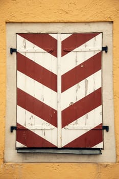Aged wooden shutters with red and white diagonal stripes on a textured yellow wall. The peeling paint and vintage metal hinges add a rustic, historical character
