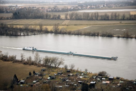 Cargo barge sailing on a wide river, aerial view. Rural landscape with fields and small houses in the background. Ideal for transportation, logistics, and environmental themes