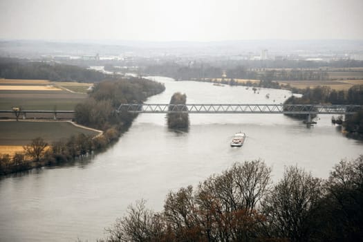 A cargo ship moving along a winding river, crossing under a steel bridge. The mix of farmland and distant city buildings adds depth, making it perfect for transport, trade, and infrastructure visuals