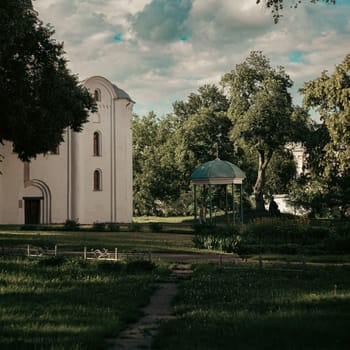 This scene shows a white church building with arched windows in a well-kept garden. A small gazebo is visible in the center of the garden, shaded by tall trees.