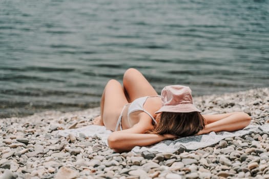 A woman is laying on the beach with a pink hat on her head. She is wearing a white bikini and is laying on a towel. The beach is rocky and the water is calm. Scene is relaxed and peaceful