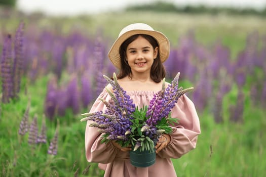 Young caucasian female child holding lupine flowers in field, wearing straw hat.