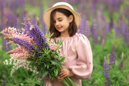 Young caucasian girl in sun hat holding colorful lupine flowers in blooming field.