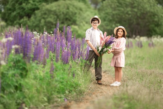 Young caucasian boy and girl in field with purple lupine flowers, smiling and holding bouquets.