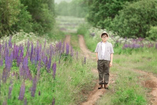 Caucasian male child in hat standing on forest path with purple wildflowers.