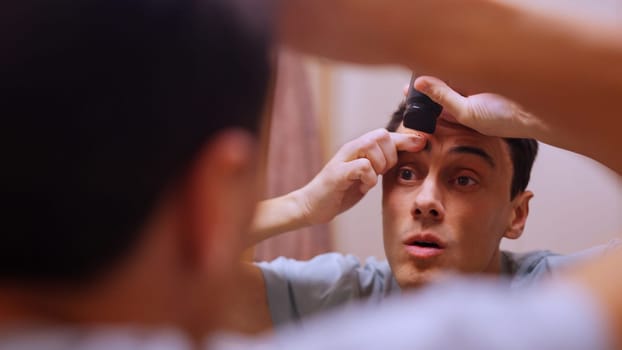 Caucasian man trimming his eyebrows with an electric razor, looking in the bathroom mirror