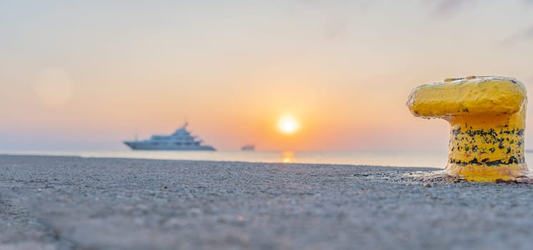 Bright yellow bollard on pier on Paradiso Beach with defocused luxury boat moored on Kos coastline with Blue Star Ferry in background on Greek Island.