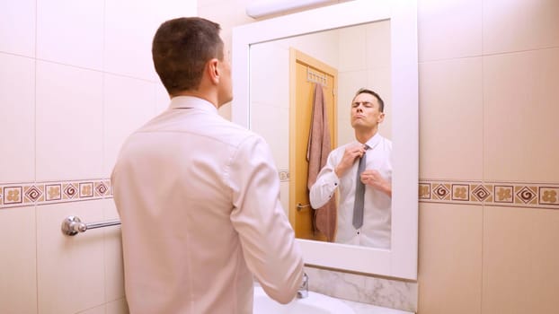 Elegant successful businessman adjusting his tie in front of a mirror in the bathroom, preparing for the workday