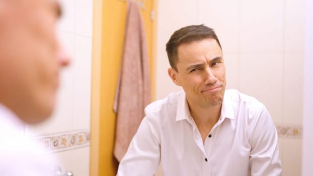 Businessman wearing white shirt making funny faces and looking at himself in the bathroom mirror