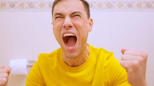 Man with a yellow t shirt is screaming and celebrating his victory while sitting on the toilet
