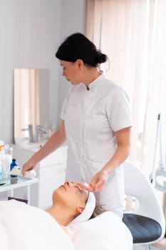 Dermatologist taking product in hand for female client lying on massage table at salon. Relaxed young woman is undergoing facial treatment in beauty spa