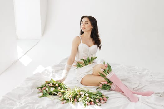 Elegant woman in lingerie lying on a bed with fresh tulips, wearing sheer gloves and pink socks, in a bright, minimalistic interior
