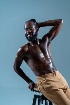 Happy shirtless black gay model leans casually on stool against blue background, showcasing confidence and joy with warm smile, exuding sense of positivity and self-expression