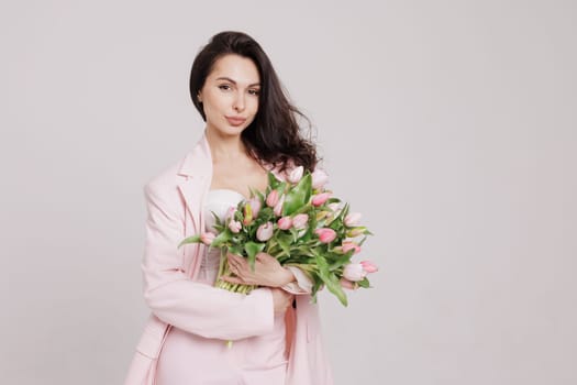 Smiling woman in a pink suit holding a bouquet of fresh tulips, posing against a neutral background in a soft, feminine, and elegant spring-inspired look