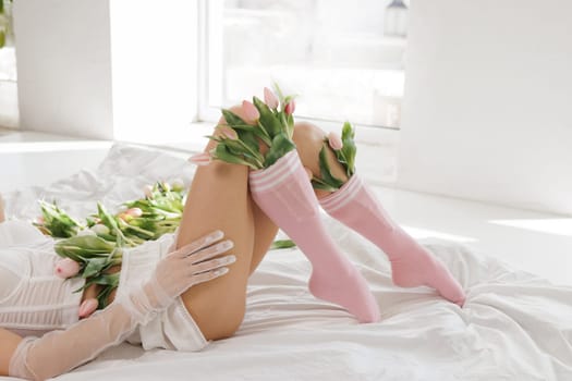 Elegant woman in lingerie lying on a bed with fresh tulips, wearing sheer gloves and pink socks, in a bright, minimalistic interior