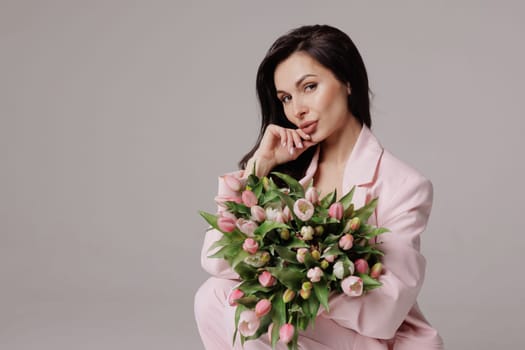 Smiling woman in a pink suit holding a bouquet of fresh tulips, posing against a neutral background in a soft, feminine, and elegant spring-inspired look