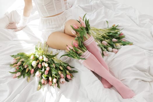 Close-up of a woman's legs in pink socks with fresh tulips, sitting on a white bed in soft natural light, creating a dreamy, romantic spring mood.