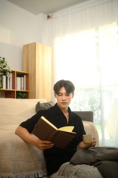 Young asian man reading a book while drinking hot drink in bright living room.