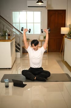 Young man exercising with dumbbells and following a workout tutorial on digital tablet.