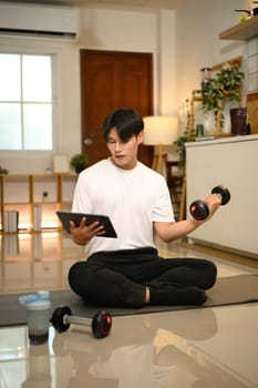 Young man lifting a dumbbell while following a workout tutorial on digital tablet.
