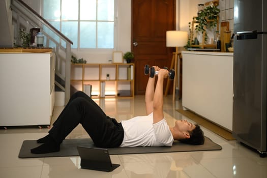 Young man lifting dumbbells on exercise mat and following an online workout session on digital tablet.