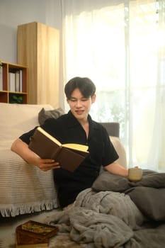 Young asian man reading a book while drinking hot drink in bright living room.