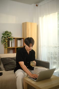 Young asian man holding a coffee cup and working on his laptop in a living room.