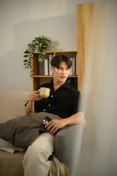 Young man holding a coffee cup sitting on couch and looking away.