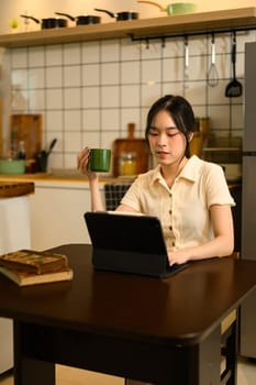 Happy woman sitting at the dining table sipping coffee and using digital tablet.