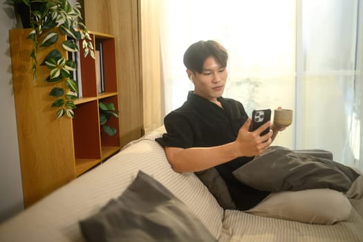 Happy young man wearing wireless earphones and using smartphone while enjoying a cup of coffee.