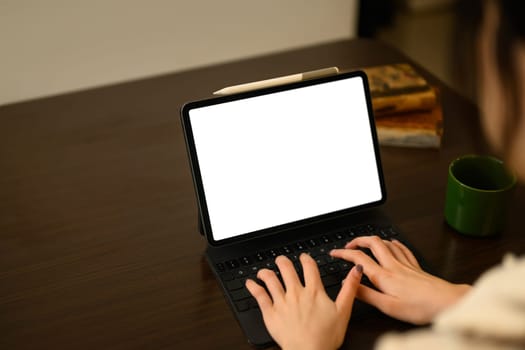 Close up shot of a woman hands typing on digital tablet with keyboard on a wooden dining table.