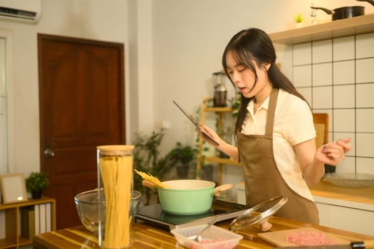 Pretty young woman in apron following a recipe on digital tablet while cooking spaghetti in a kitchen.