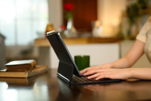 Close up shot of a woman hands typing on digital tablet with keyboard on a wooden dining table.