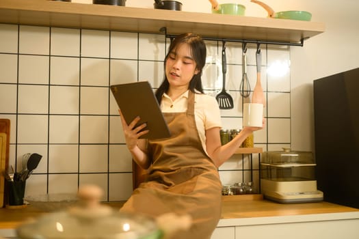 Young woman sitting on the kitchen counter and checking a recipe on digital tablet.