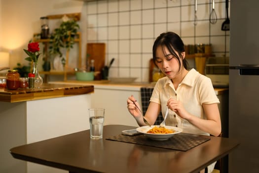 Pretty asian woman sitting at a dining table, enjoying a cozy meal in a warm kitchen.
